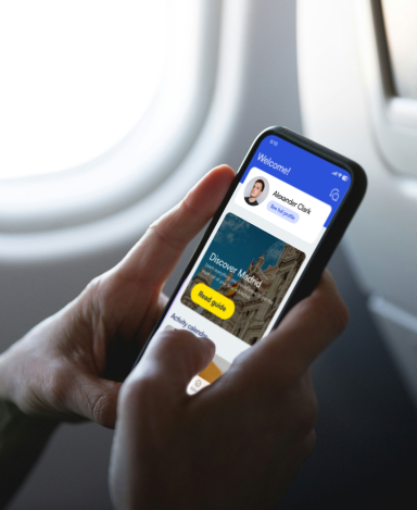 Pasajero usando la aplicación móvil Lodgerin a bordo de un avión, con pantalla de bienvenida personalizada para 'Ricardo Díaz' y una sección 'Discover Madrid' con botón amarillo. Smartphone mostrado junto a ventanilla de aeronave durante vuelo.